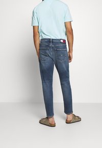 Man som bär en ljusblå T-shirt, blå Tommy Jeans och bruna slip-on sandaler, stående vänd mot en enkel vit vägg.