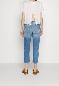 Vit kortärmad topp med hålmönster och öppning i ryggen, kombinerad med ljusblå jeans i ankellängd och bruna slip-on skor.