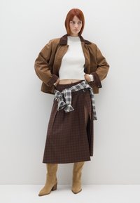 Braune Baumwolljacke mit Steppkragen, weißes geripptes Crop-Top, brauner karierter Rock mit Seitenschlitz und beige High Heels. Schwarz-weißer karierten Krawatte.