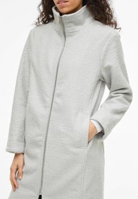 Pelēks vilnas mētelis ar augstu apkakli, priekšējo rāvējslēdzēju slēgumu un sānu kabatām. Audums ir ar teksturētu virsmu un vaļīgu piegriezumu.