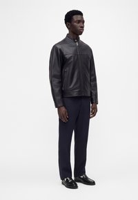 Jeune homme debout portant une veste zippée en cuir noir, un pantalon foncé et des chaussures habillées noires et brillantes sur un fond blanc uni.