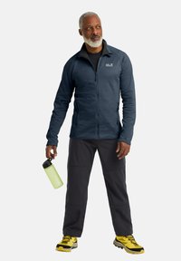 Giacca a zip da uomo di colore blu scuro, abbinata a pantaloni neri e scarponcini da trekking gialli. Tiene in mano una borraccia verde, in piedi davanti a uno sfondo neutro.