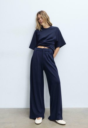 Femme debout contre un mur blanc, portant un pantalon large bleu marine, un haut à manches courtes assorti et des chaussures plates blanches, avec une main dans la poche.