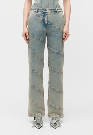 Woman wearing light blue denim pants with diagonal seam details and silver pointed-toe heels with bow accents, standing against white background.