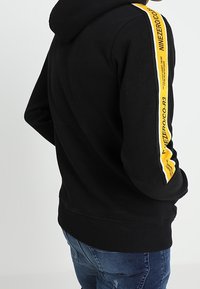 Felpa nera con una striscia logo gialla lungo la manica sinistra. Presenta una vestibilità classica e un cappuccio regolabile con cordini. Materiale in misto cotone.