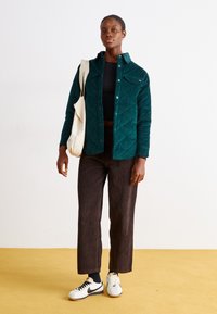 Veste en velours côtelé teal avec un motif en losanges, crop top noir, pantalon en velours côtelé marron à jambes larges et baskets blanches avec accents noirs.