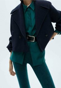 Giacca cropped blu scuro con doppi bottoni, abbinata a una camicia verde con bottoni e leggings turchesi, caratterizzati da texture e styling a strati.