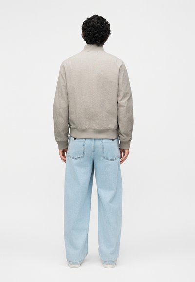 Homme aux cheveux bouclés, debout de dos, portant une veste beige texturée et un jean bleu clair ample sur un fond uni.