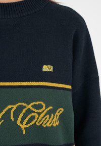Granatowy sweter z prążkowanym kołnierzem, z żółtymi i zielonymi poziomymi paskami oraz haftowanym napisem "Klub" w kursywie.