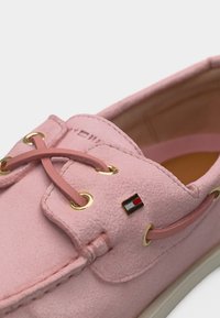 Ροζ μοκασίνι από σουέτ με χρυσά μεταλλικά τρύπια κορδονιών, ροζ κορδόνια στο ίδιο χρώμα και ένα μικρό ορθογώνιο εμβληματικό σημαιάκι στο πλάι κοντά στα κορδόνια.