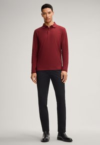 Polo a maniche lunghe di colore bordeaux con un motivo a diamante, abbinato a pantaloni neri e stivali neri, su uno sfondo neutro.