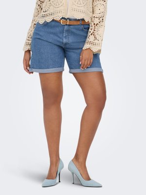Femeie purtând pantaloni scurți din denim albastru rulati, o curea maro deschis, un top bej croșetat și pantofi cu toc înalt bleu, stând în fața unui fundal simplu.