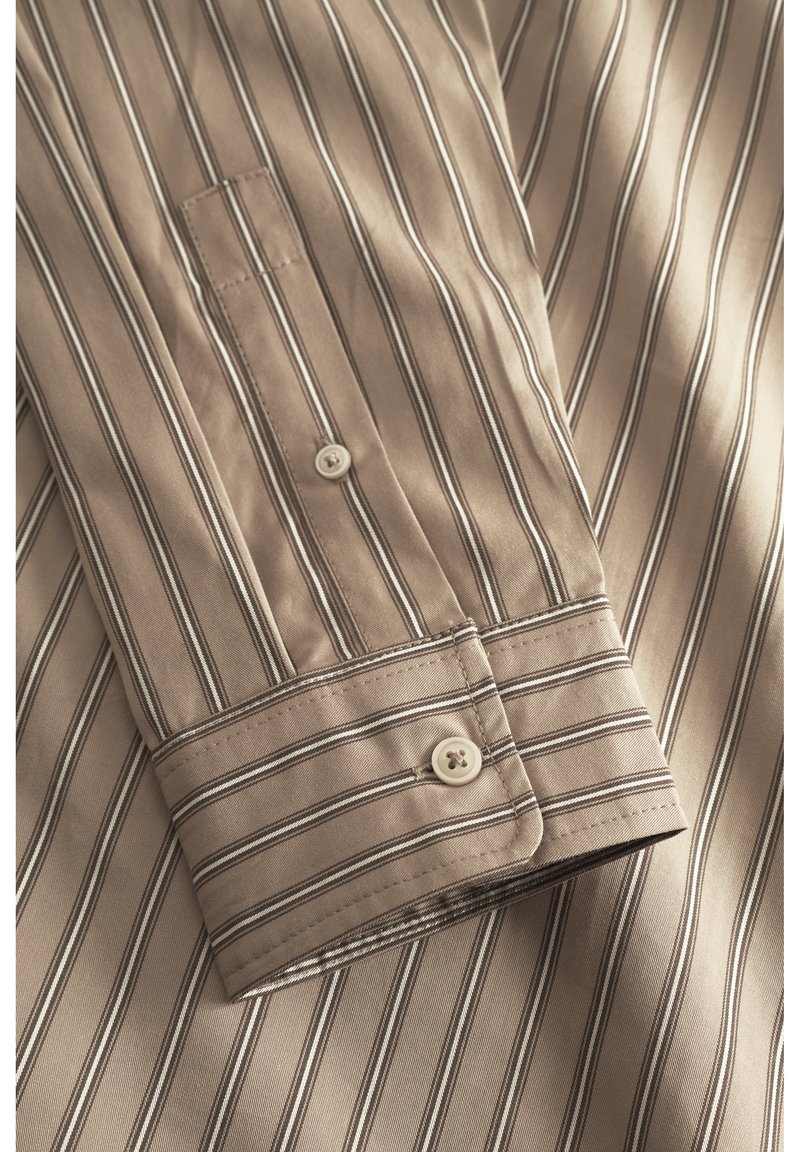 Camicia beige a maniche lunghe con sottili strisce verticali marroni, dotata di polsino abbottonato e taschino sul petto. Tessuto di cotone liscio.