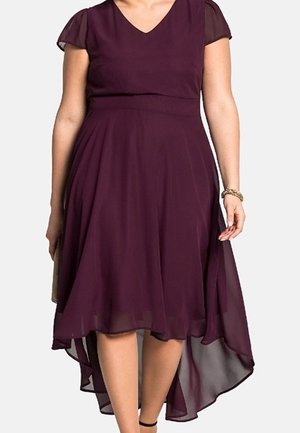 Kvinde iført en ærmeløs, dyb lilla high-low kjole med V-hals og gennemsigtige kappelærmer, accessoriseret med armbånd og clutch.