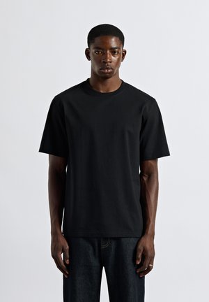 Young man stands facing forward wearing a plain black short-sleeve t-shirt and dark denim jeans against a light background.