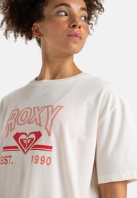 T-shirt couleur crème avec le logo "ROXY" rouge et un design de cœur, encolure ronde et manches courtes en tissu de coton doux.