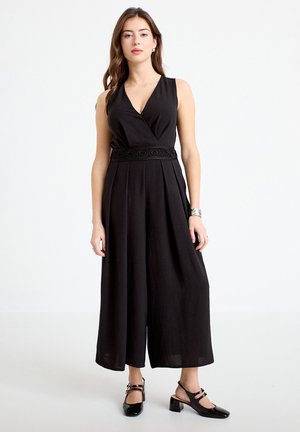 Kobieta w czarnym bezrękawnym kombinezonie z szerokimi nogawkami i ozdobnym pasem w talii, w połączeniu z czarnymi butami na słupku.