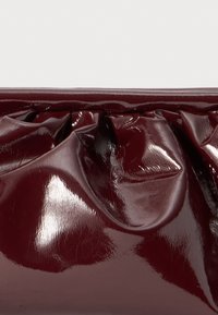 Viininpunainen clutch-laukku, jossa on kiiltävä, ryppyinen pinta. Laukussa on rypytetty muoto ylhäällä ja sileä viimeistely. Kompakti suorakulmainen muoto.
