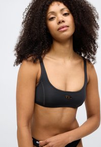Juoda sportinė liemenėlė su apvalia iškirpte, storomis petnešėlėmis ir logotipo akcentu. Lygi tekstūra ir elastinis audinys suteikia komfortą ir palaikymą.