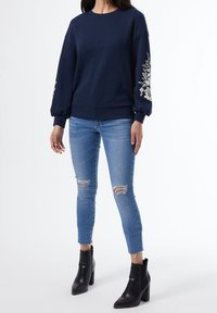 Mörkblå sweatshirt med blommig broderi på ärmarna, matchad med ljusblå slitna jeans och svarta ankelstövlar med klack.