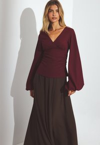 Burgundska majica z dolgimi rokavi z teksturirano tkanino in V-izrezom, v kombinaciji z lahkotno rjavo krilo. Gladka površina, eleganten kroj, brez vzorcev.