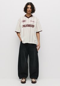 Weißes Netz-Jersey mit weinroten Streifen, dem Text "MADRID" und dekorativen Mustern. Kombiniert mit lockeren schwarzen Hosen und braunen Schuhen.