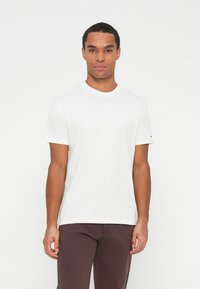 Joven de pie, vistiendo una camiseta blanca de manga corta y pantalones marrón oscuro contra un fondo blanco.