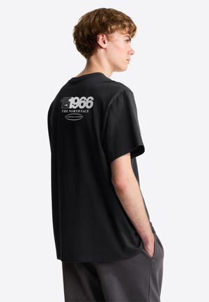 Młody mężczyzna w czarnej koszulce z logo "1966 THE NORTH FACE" na plecach, stoi z rękami w kieszeniach, zwrócony w prawo na białym tle.