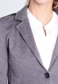 Cárdigan gris con un diseño ajustado, que cuenta con un cierre de botones, un cuello de muesca y acentos texturizados en los hombros.