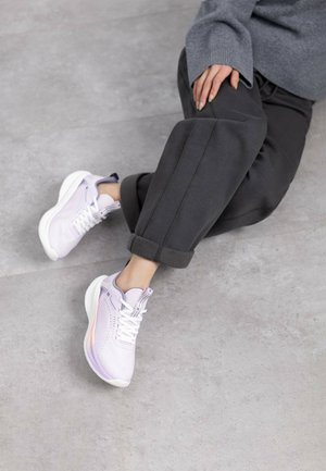 Zapatillas deportivas de color púrpura claro con suelas blancas, parte superior de malla y cierre de cordones. Combinadas con joggers oscuros de gris con puños sobre una superficie texturizada.