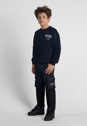 Fekete pulóver felirattal, hosszú ujjak, kerek nyakú; fekete cargo nadrággal párosítva, amely foltokkal és hímzett részletekkel rendelkezik; sötét sportcipő.