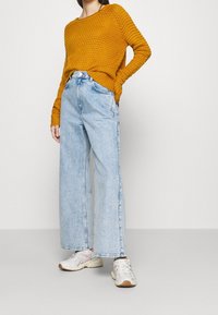 En person som bär en senapsgul stickad tröja, vida ljusblå jeans och vita sneakers, står framför en neutral bakgrund.