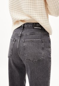 Personne portant un jean noir délavé taille haute et un pull en tricot crème, montrant les poches arrière et l'étiquette de marque sur la ceinture.