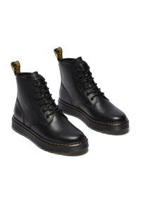 Botas de cuero negro hasta el tobillo con punta redonda, cordones en la parte delantera y una pestaña amarilla. Presentan una suela de goma gruesa con costuras visibles.