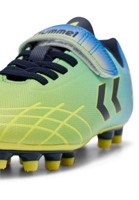 Chaussure de football synthétique verte et bleue avec accents noirs, surface texturée, motif en grille jaune et semelle extérieure à crampons noirs.