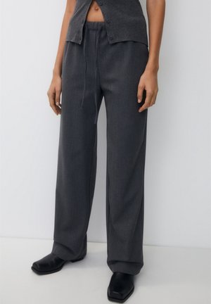 Personne portant un pantalon ample gris foncé à cordon, des chaussures noires à bout carré et un haut sombre boutonné sur fond blanc.