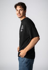 Schwarzes Baumwoll-T-Shirt mit kurzen Ärmeln und rundem Hals, mit einer weißen Grafik auf der linken Brust. Kombiniert mit blauen Jeans.