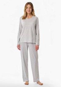 Nő mezítláb áll, világosszürke csíkos hosszú ujjú felsőt és laza szürke pizsamát visel, fehér háttér előtt.