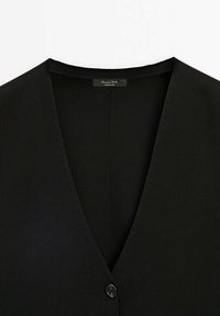 Cardigan noir à col en V avec un bouton visible et une étiquette à l'intérieur du col indiquant la marque et fabriqué en Chine.