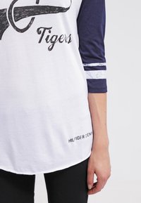 Valkoinen T-paita, jossa tummansiniset 3/4-hihat, mustalla painatuksella sana "Tigers" ja valkoiset raidat hihoissa. Merkitty tekstillä "HILFIGER DENIM."