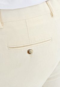 Jasno beżowe lniane spodnie z tylną kieszenią ozdobioną dwoma poziomymi paskami oraz okrągłym, jasnobrązowym zapięciem na guzik.