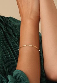 Goldarmband mit länglichen rechteckigen Gliedern und kleinen runden Akzenten, gezeigt an einem Arm vor einem strukturierten grünen Stoffhintergrund.