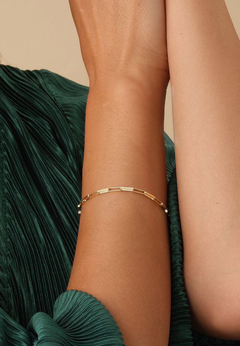 Goldarmband mit länglichen rechteckigen Gliedern und kleinen runden Akzenten, gezeigt an einem Arm vor einem strukturierten grünen Stoffhintergrund.