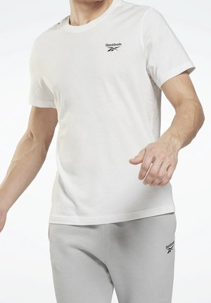 Mężczyzna w białej koszulce Reebok i jasnoszarych spodniach dresowych z logo Reebok, stojący z lekko zgiętymi ramionami.