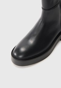 Černá kožená kotníková bota s hladkým povrchem, kulatou špičkou a pevným gumovým podešví. Jednoduché prošívání zvýrazňuje design.