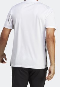 Bílé sportovní tričko s krátkými rukávy, hladkou látkou a síťovanými panely po stranách. Černé pruhy vedou svisle pod él stran.