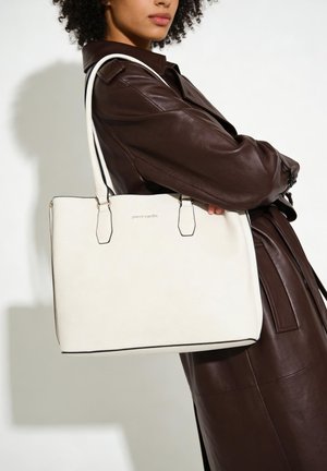 Personne portant un manteau en cuir marron foncé tient un grand sac cabas blanc Pierre Cardin sur l'épaule.