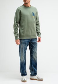 Groene sweatshirt met ronde halslijn en logodetails. Gedragen met blauwe versleten jeans en witte sneakers met grijze accenten.
