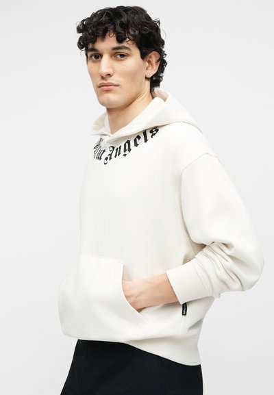 Jeune homme aux cheveux bouclés foncés portant un sweat à capuche blanc avec un texte noir près du col et un pantalon noir, les mains dans les poches avant.