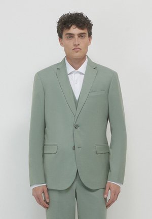 Abito verde chiaro in tessuto strutturato, con chiusura a due bottoni, revers a punta e due tasche frontali. Indossato sopra una camicia bianca.